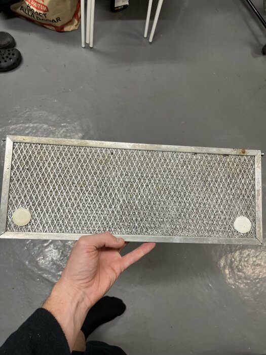 En hand håller en metallfilterpanel över ett grått golv, bakgrunden visar suddiga föremål och skor.
