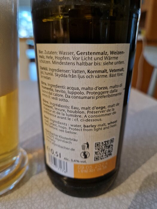 Ölflaska med ingredienser på flera språk, datumstämpel, delvis synligt ölglas i bakgrunden.