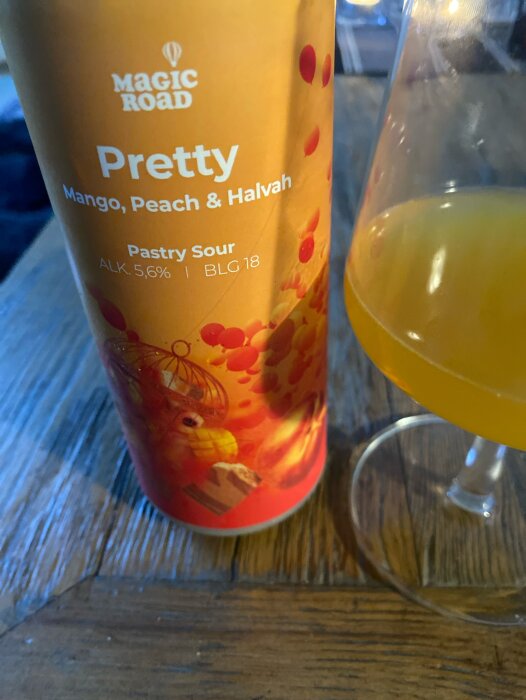 En ölburk med texten "Pretty Mango, Peach & Halvah", ett glas med dryck, träyta.