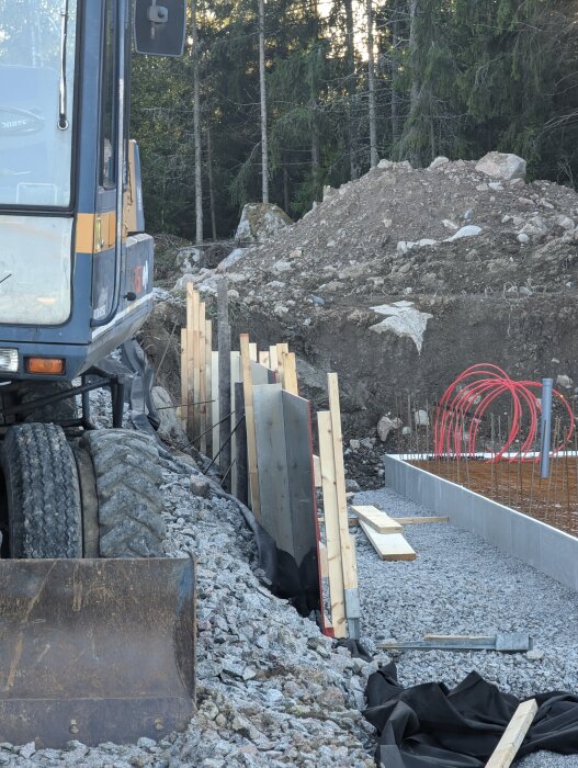 Grävmaskin vid byggarbetsplats med betongfundament, skog i bakgrunden, rör och konstruktionsmaterial synligt.