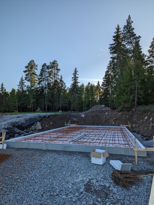 Grundläggning för byggnad, armeringsjärn och värmeslingor på grus, skogsbakgrund, blå himmel.