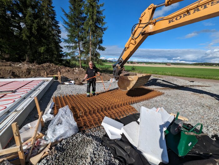 Arbetsplats utomhus med grävmaskin, arbetare, förstärkningsnät och konstruktionsmaterial för grundläggning. Lantlig bakgrund med träd.