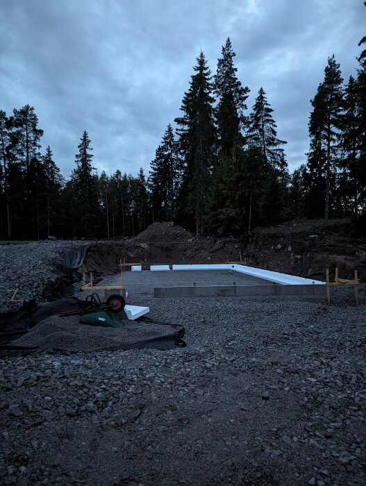 Byggarbetsplats i skymningen, grundkonstruktion, grus, skogsbakgrund, molnig himmel, övergivet byggmaterial.