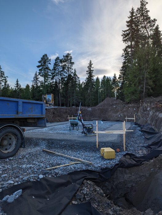 Byggarbetsplats med grus, lastbil, utsattsutrustning, skogsbakgrund, tillfällig träkonstruktion, jordhög.