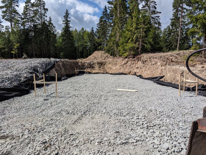 Grundläggning av anläggning eller bygge, grus, svart markduk, skogsbakgrund, molnig himmel.