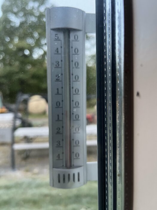 Utomhustermometer monterad på ett fönster, visar cirka 4 grader Celsius, suddig bakgrund med träd och gräs.