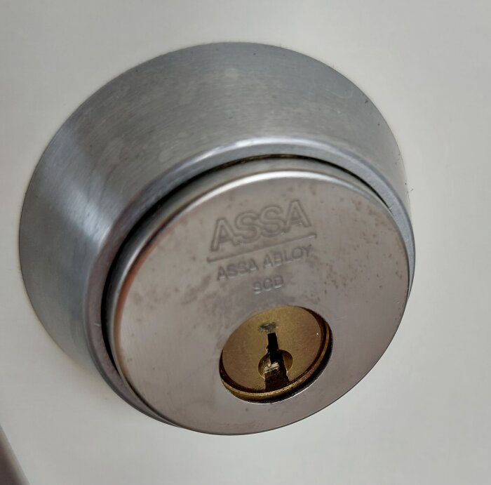 ASSA-cylinderlås, nyckelhål, metall, säkerhetsenhet, ASSA ABLOY-logotyp, skruvar, vit bakgrund.