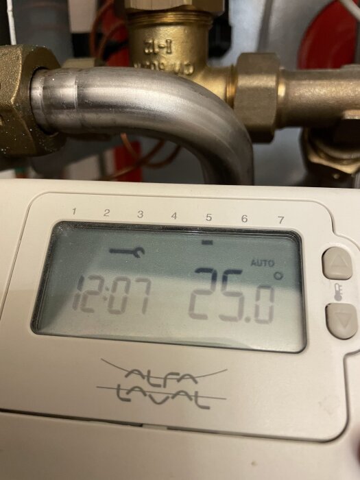 Termostat med digital display, visar 25.0 grader Celsius och tiden 12:07, rör och ventiler i bakgrunden.