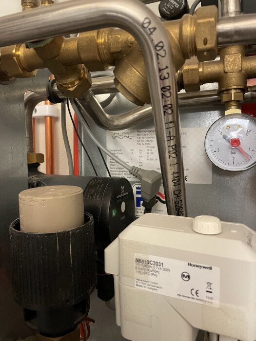 VVS-installation med mässingsrör, tryckmätare, cirkulationspump och Honeywell-komponent i en teknisk anordning.