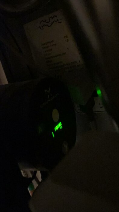 Mörk bild visar elektronisk enhet med gröna strömindikatorljus, oskarp text och etiketter i bakgrunden.