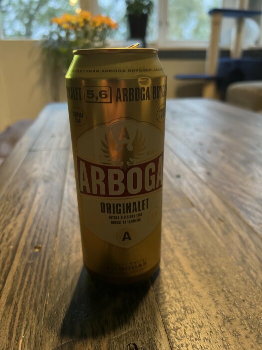 En ölburk av märket Arboga står på ett träbord, inomhus med suddig bakgrund och dagsljus.