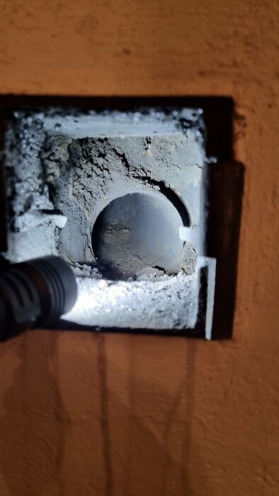 En ficklampa lyser på en isolerad vägggenomföring synlig genom en rektangulär öppning i väggen.
