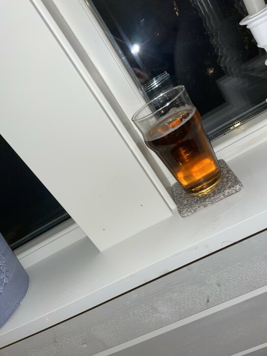 Ett glas med bärnstensfärgad dryck på fönsterbrädan om kvällen, reflektion och sned vinkel.