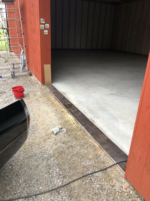 Ett tomt garage med betonggolv, röda väggar, en stege, en röd hink och tidningspapper på marken.