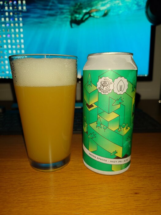 Ölglas med skum bredvid ölburk, "Green Spaces Hazy IPA", datorskärm med simmare i bakgrunden.