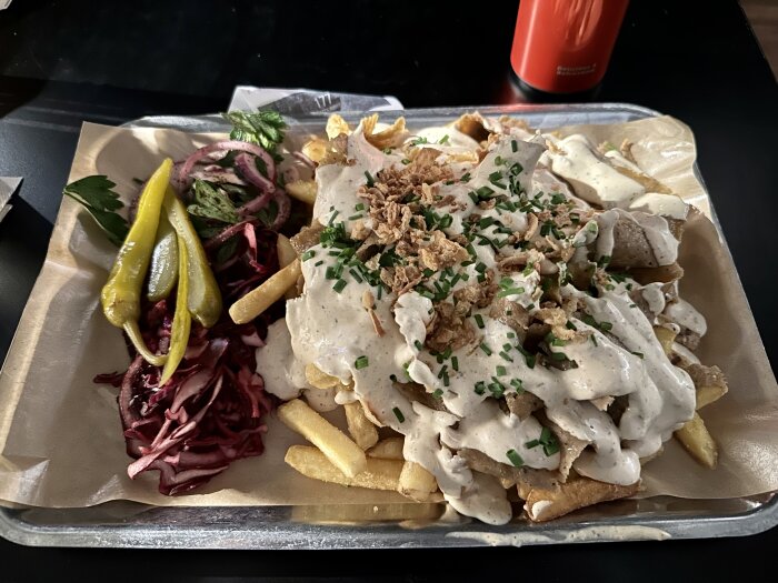 Pommes frites med kebabkött, sås, inlagda grönsaker, rödlök, persilja på silverbricka, orange dryckesflaska bakom.