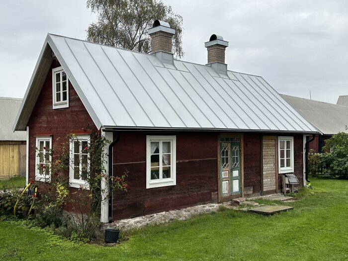 Rött trähus, vita fönster, plåttak, skorstenar, rosbuskar, grön gräsmatta, landsbygd, molnig himmel.