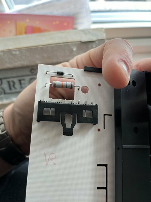 En hand håller en vit elektronisk kretsplatta med resistor och andra komponenter mot en otydlig bakgrund.