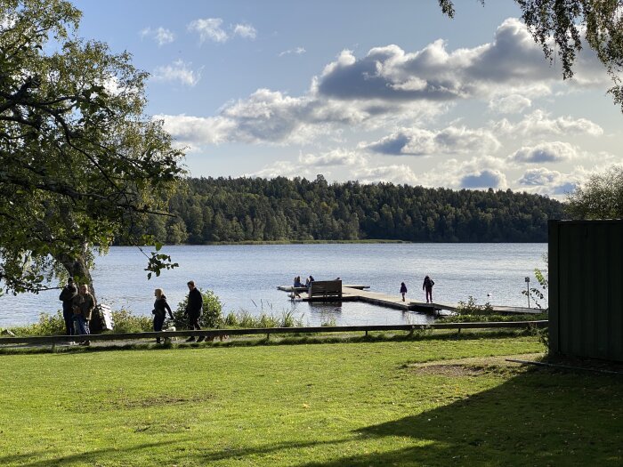 Sjöutsikt, grönområde, människor promenerar, brygga, moln, träd, soligt, avkopplande, utomhusaktivitet, vattenspegling, natur.