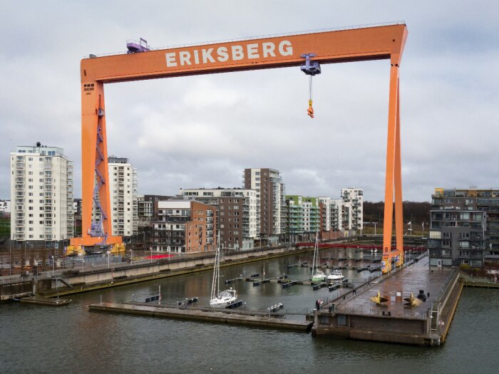 Stor orange kran märkt "ERIKSBERG", båtar och moderna byggnader vid vatten, molnig himmel.