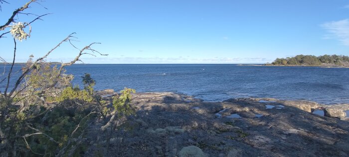 Skärgårdsmiljö, klippor, vatten, träd, blå himmel, dygnets ljusa timmar, fridfull, orörd natur.