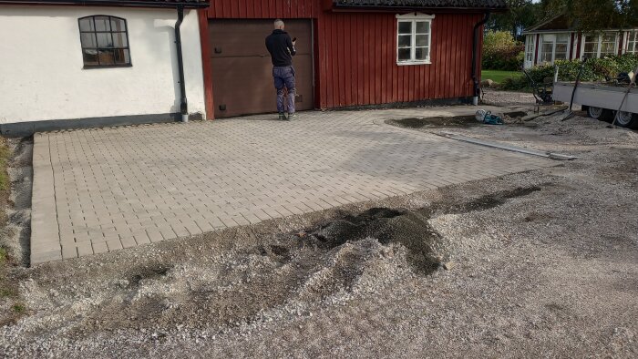 Uppfart under konstruktion, person står vid garage, marksten, husfasad, byggmaterial, trädgårdsvagn, bladlös vegetation.