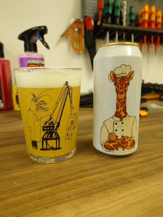 Ett glas öl med kranillustration bredvid en burk med giraffkockmotiv på ett träbord, verktygsvägg i bakgrunden.