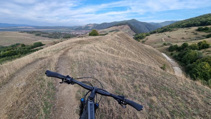 Mountainbikeperspektiv på lantlig stig, kullar, gröna träd, delvis molnig himmel, och en liten by.