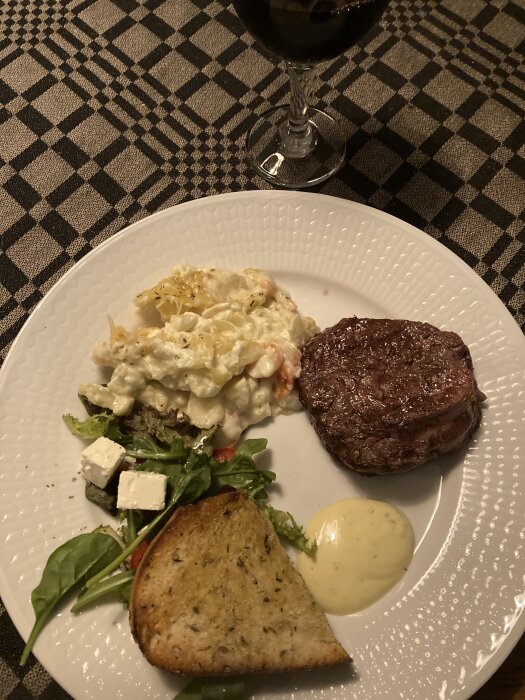 Tallrik med steak, potatissallad, grönsallad, rostat bröd, bearnaisesås, rödvin i glas, rutig duk.