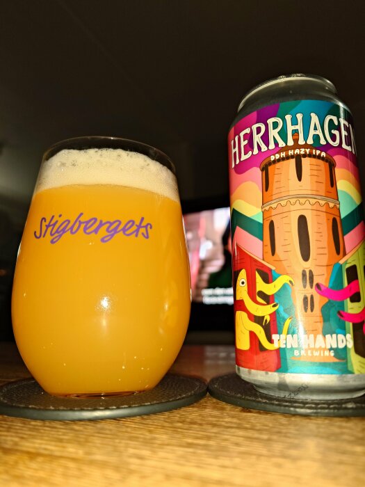 Ölglas med skum, grumlig öl bredvid färgglad burk "Herrhagen", bordsunderlägg, del av TV-skärm.