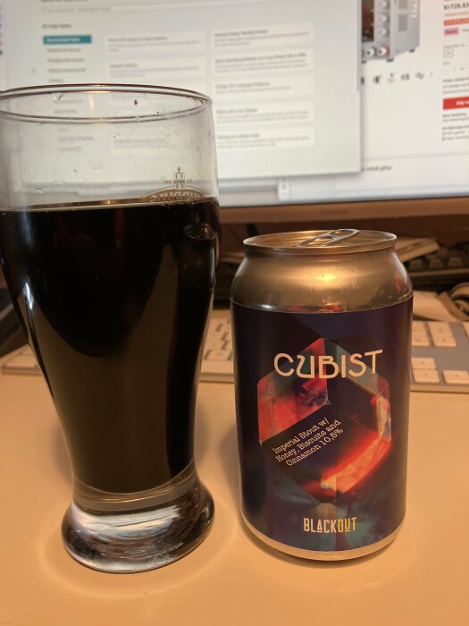 Ett glas mörkt öl bredvid en burk, "CUBIST" öletikett, kontorsmiljö, datorskärm i bakgrunden.