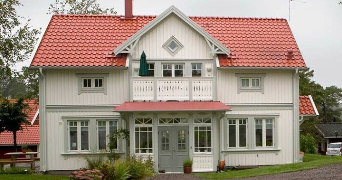 Tvåvånings trähus, vitt med rött tak, balkong, grönskande trädgård, lantlig stil.