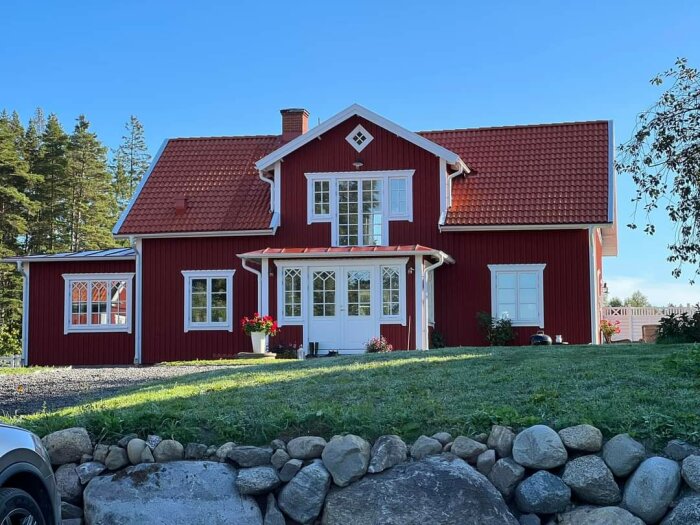 Rött trähus med vita knutar, tegeltak, grönska, stenmur, typisk svensk landsbygdskänsla.