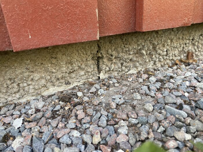 Vägghörn möter sprucken betonggrund ovanför grusig mark. Slitage eller strukturell skada synlig.