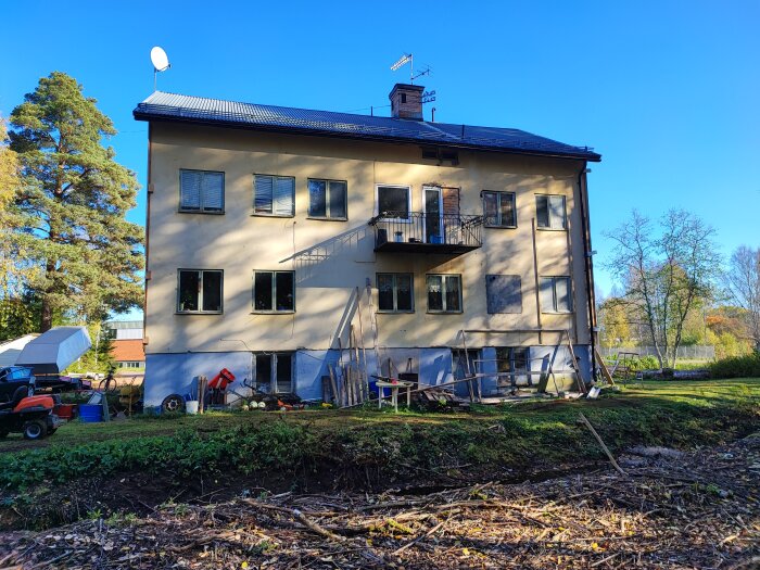 Ett gult flerfamiljshus under renovering med verktyg och material utspridda, mot bakgrund av blå himmel.