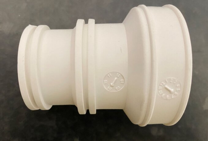 Vit plast; röranslutningsdel; olika diametrar; otydlig text; grå bakgrund.