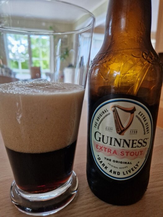 En flaska Guinness Extra Stout med ett halvfullt glas mörkt öl bredvid. Skumtäckt, inomhusmiljö.