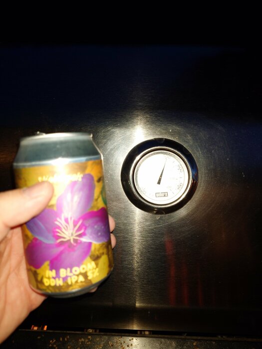 Hand håller ölburk med blomma, mot mörk bakgrund och del av termometer synlig.