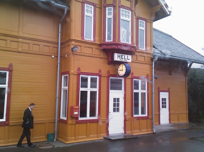 Gul träbyggnad med skylten "HELL" på fasaden, klocka, man i kostym går förbi.