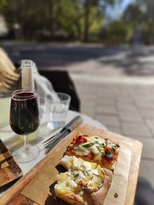 Uteservering med pizza på träbräda och ett glas rött vin, soligt väder, suddig stadsmiljö i bakgrunden.