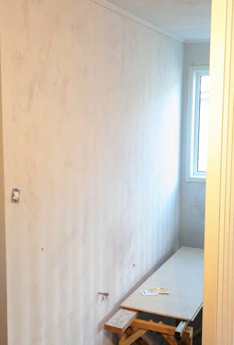 Tomt rum under renovering, vit vägg med fläckar, fönster, oskyddad elinstallation, träpall, byggmaterial.