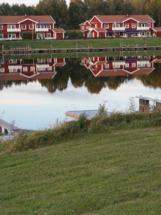 Röda hus speglas i vatten vid lugn sjö med grön gräsmatta i förgrunden.