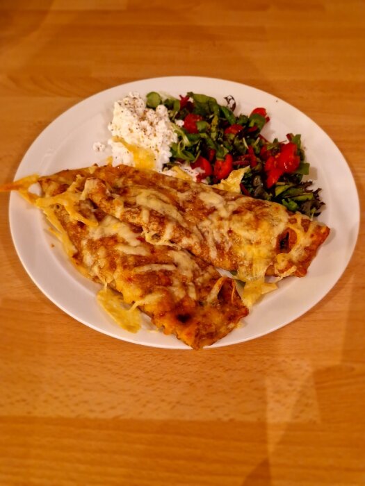 En tallrik med enchiladas täckta med ost, serverade med sallad och keso. Warm färgton. Hemlagad måltid.