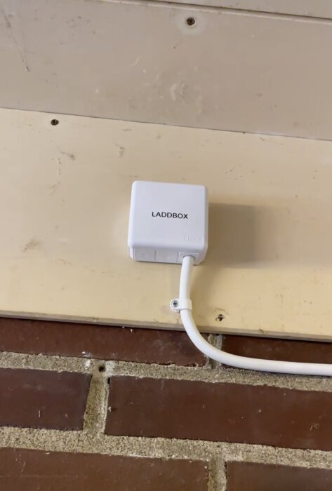 En laddbox för elbil monterad på vägg ovanför tegelsten, kabel ansluten, text "LADDBOX" synlig.