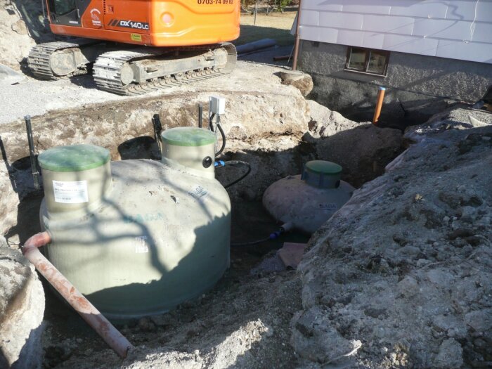Byggarbetsplats med grävmaskin, grävd grop och två installerade betongbrunnar. Husfasad och rör i bakgrunden.