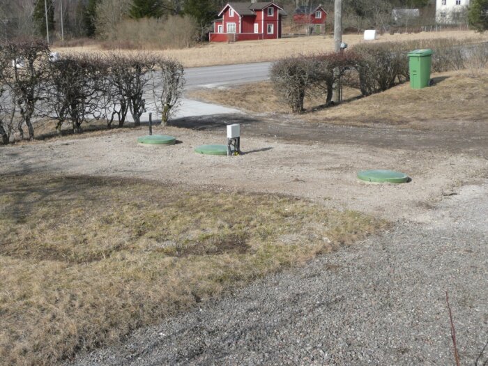 Lantlig scen, grusuppfart, röda hus, gröna brunnlock, vårljus, bar vegetation, sopkärl.