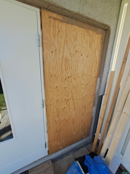 En plywoodskiva täcker en dörröppning bredvid en vit ytterdörr; renovering eller temporär lösning.