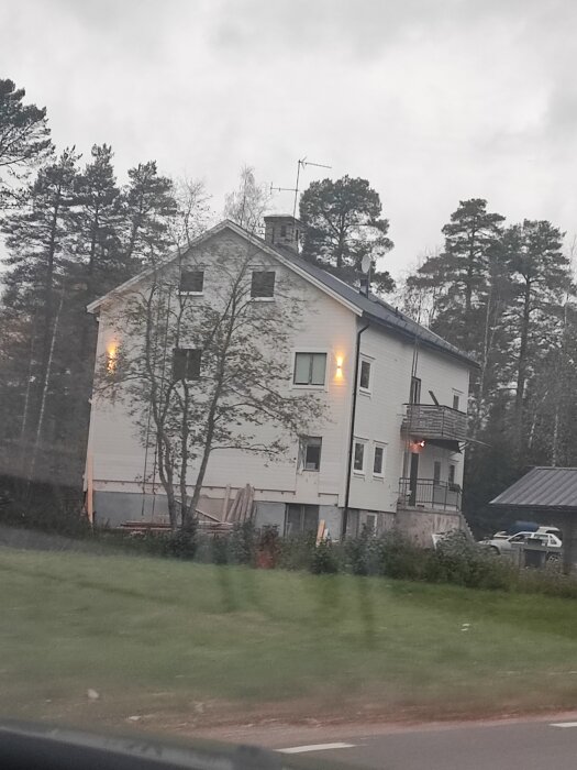 Vitt hus med tre våningar, träd, grå himmel, tagen genom fönster, suddig förgrund.