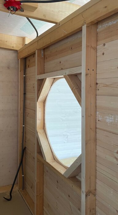 Träkonstruktion inomhus med ovalt fönster, snötäckt utsikt, byggprocess, verktyg, stolpe, ljus trä.
