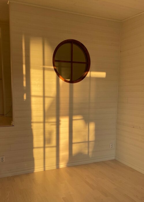 Runt fönster på trävägg, solens skuggor, varmt ljus, minimalistiskt rum, hörn, golvuttag.